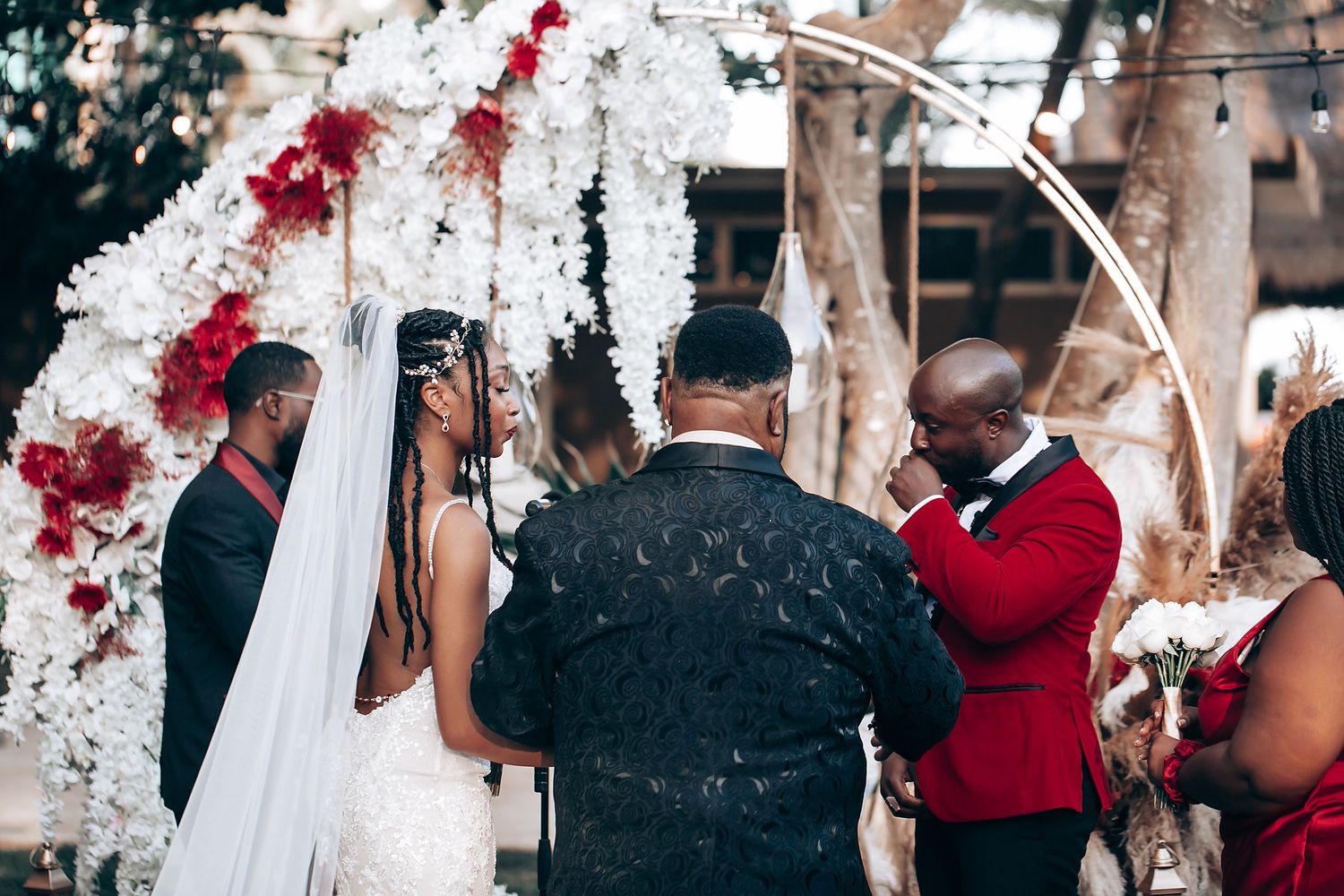 Wedding ceremony takes place under a white floral arch with red accents during an outdoor celebration.
