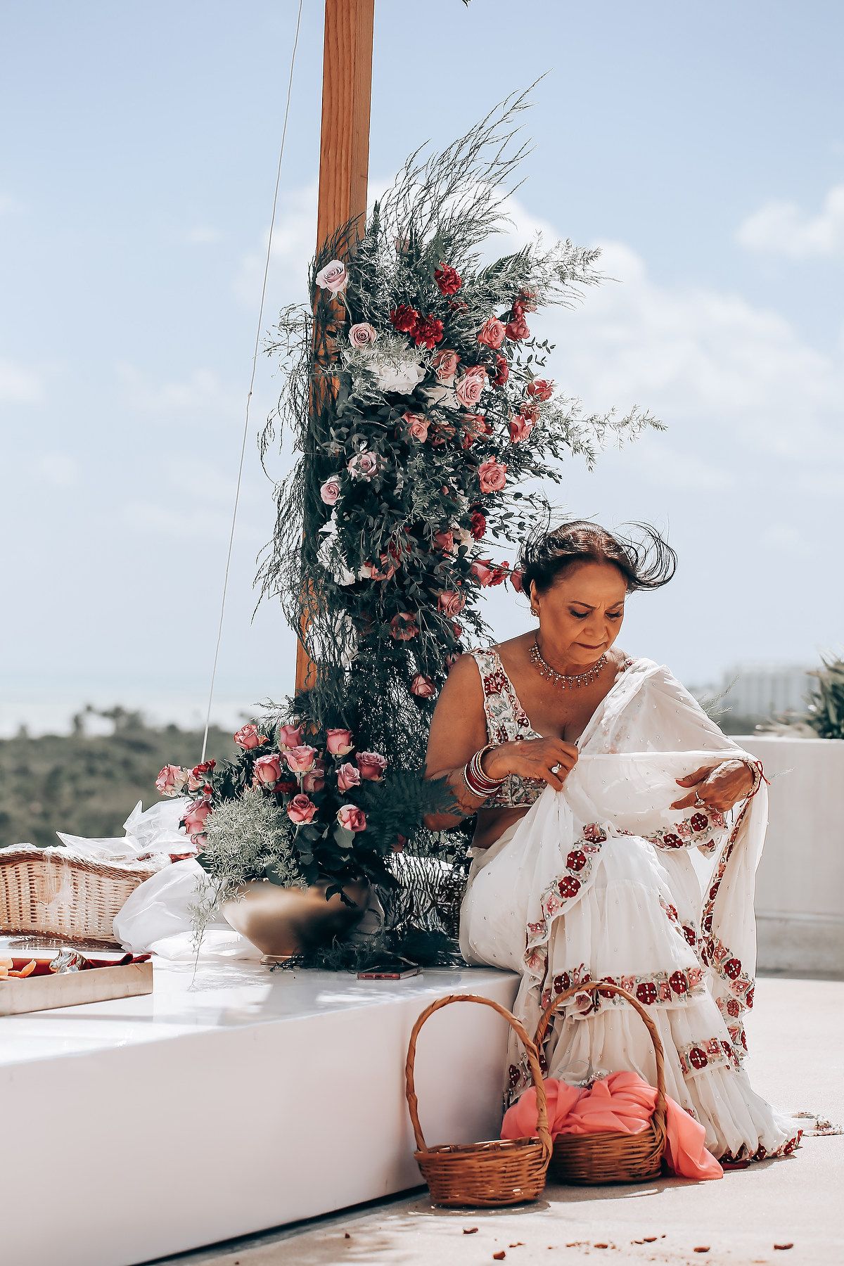 A bride in a white dress poses elegantly on a rooftop terrace with floral arrangements and pink accents.