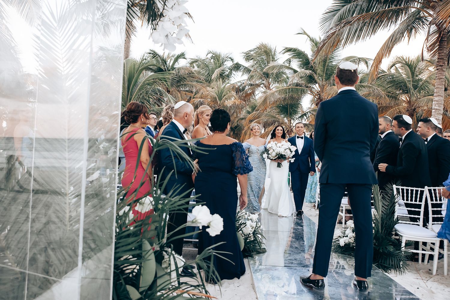 Wedding guests gather outdoors under palm trees for a tropical ceremony celebration.