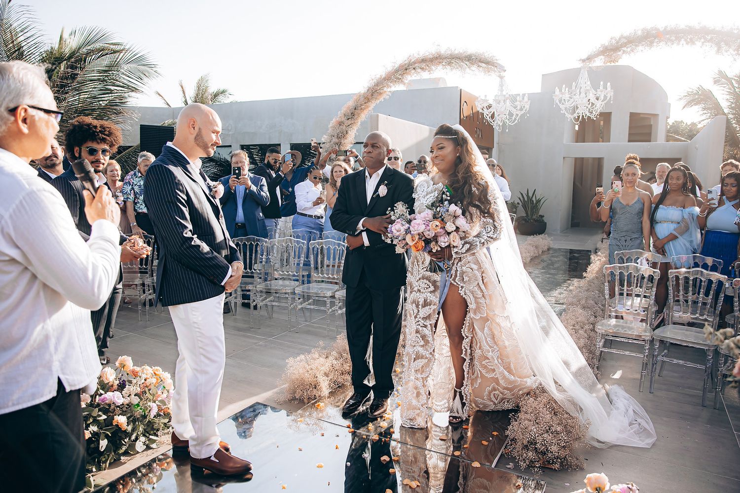 Outdoor wedding ceremony under palm trees with guests watching the celebration.