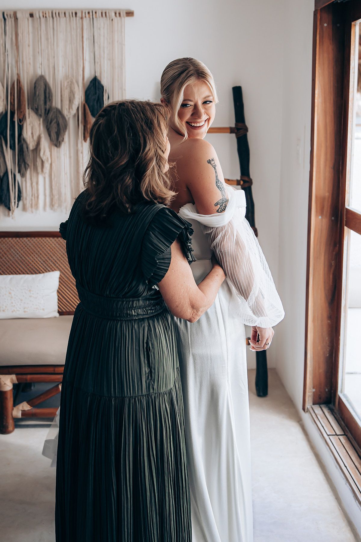 A tender moment during wedding preparations as a woman in a dark green dress helps adjust a white bridal gown.