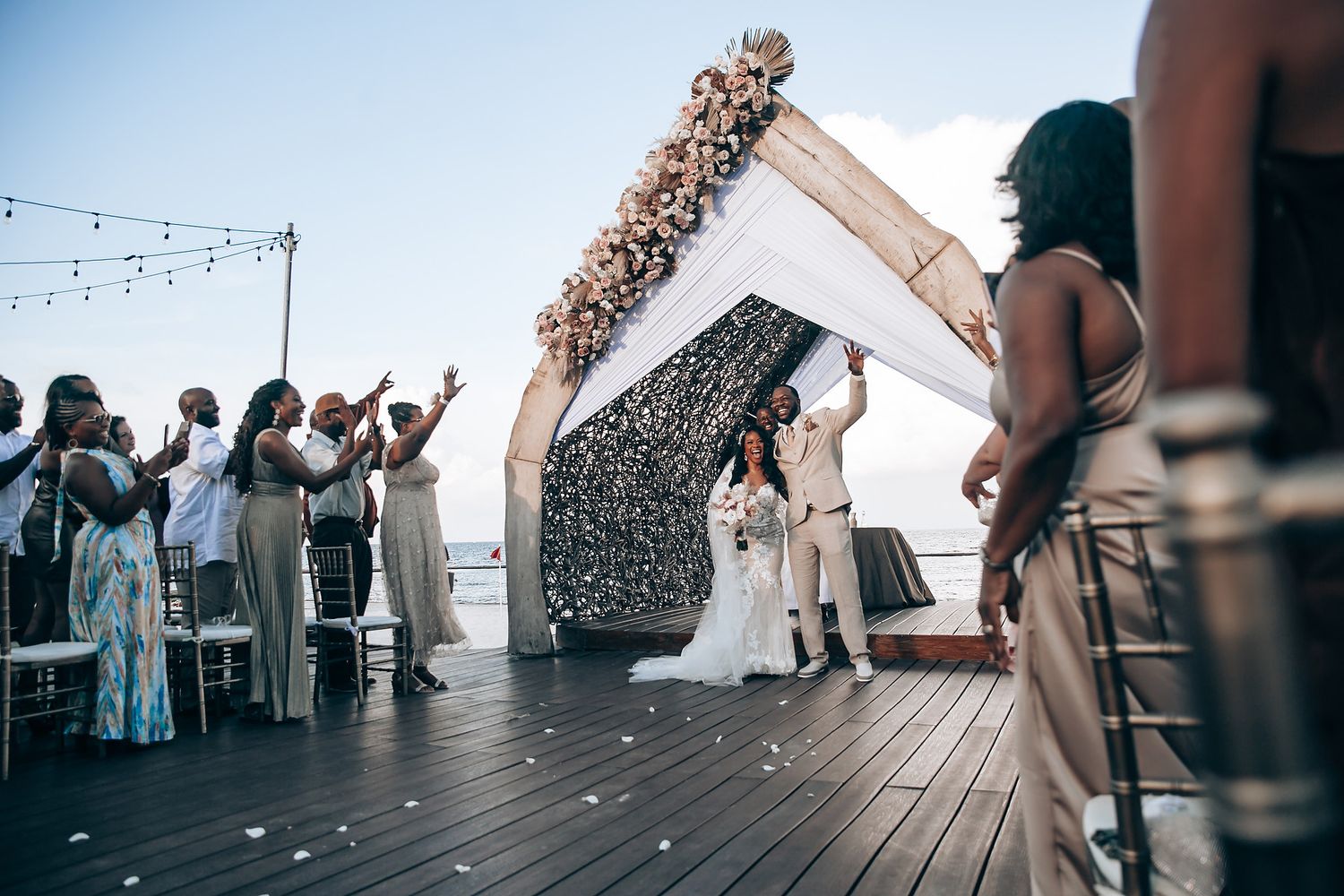 Low-angle views of wedding celebration on wooden deck with string lights overhead.