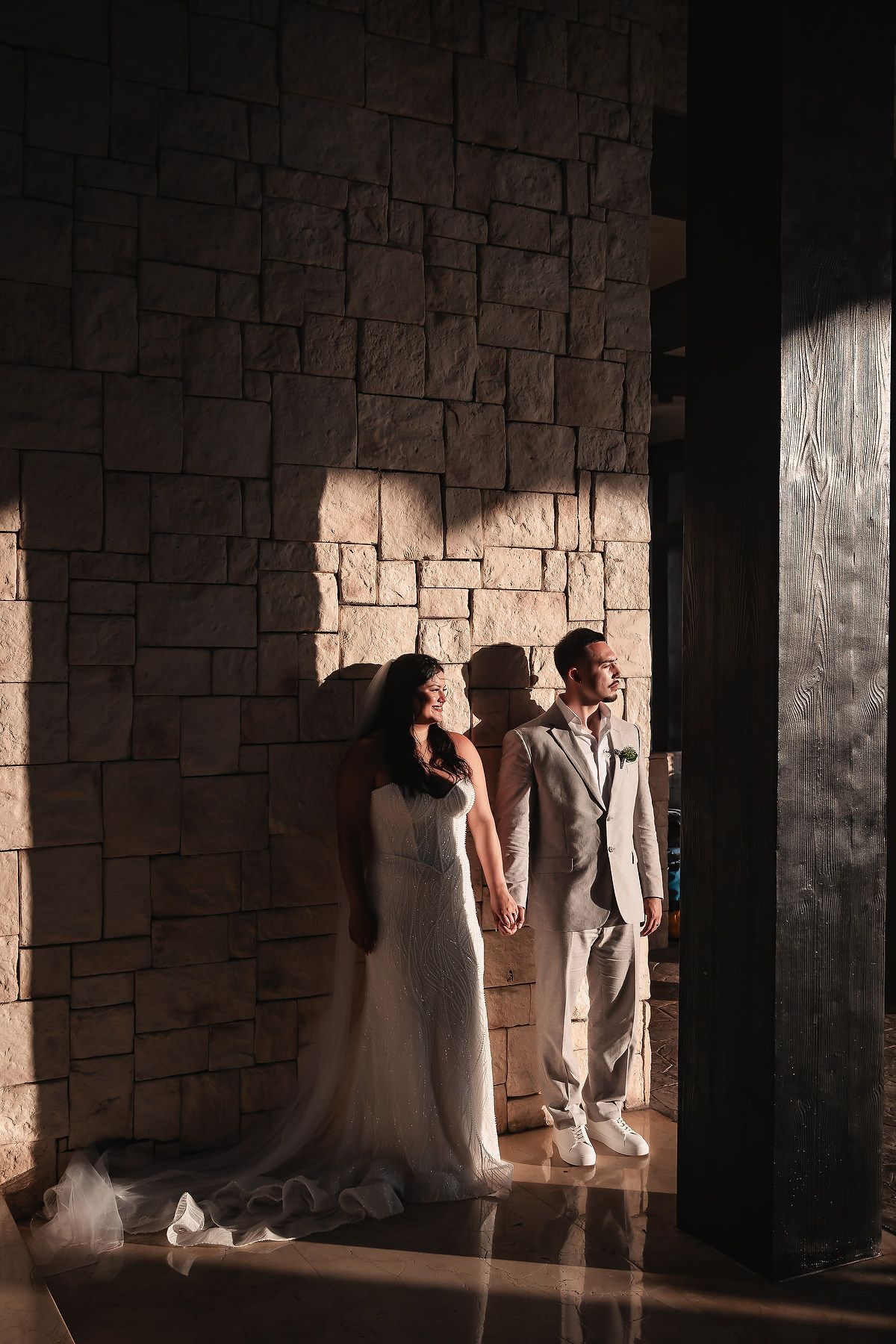 A wedding couple poses against a stone wall with dramatic lighting streaming through.