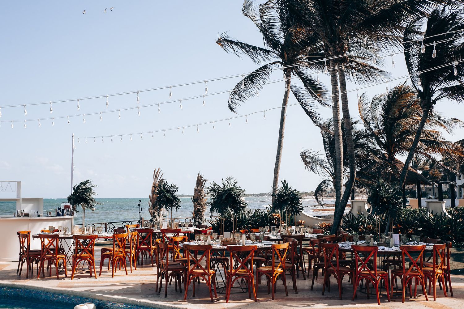 Outdoor beach reception venue with wooden tables and string lights under palm trees.