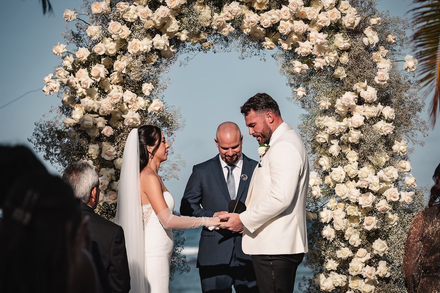 Wedding ceremony under luxurious white floral arch with tropical palm trees swaying in background.