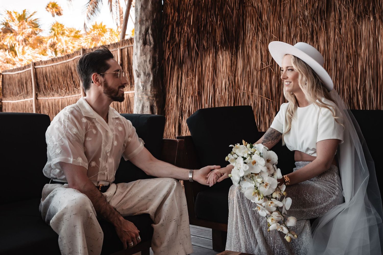 Wedding couple sitting under a rustic thatched roof setting.