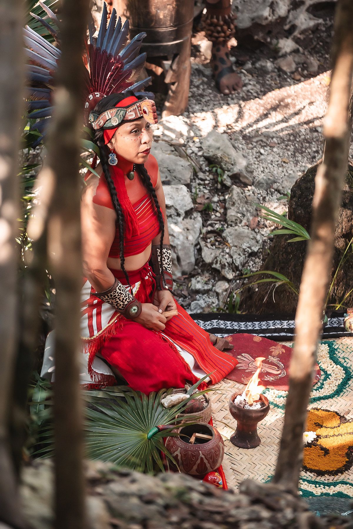 Traditional Mayan performer in red ceremonial attire sitting by ritual offerings in a cave setting.