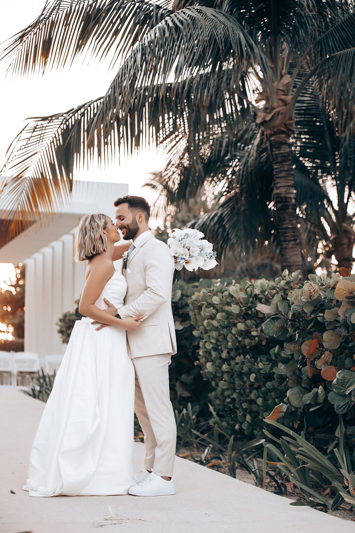 Elegant wedding couple poses against palm trees and tropical landscaping at sunset.