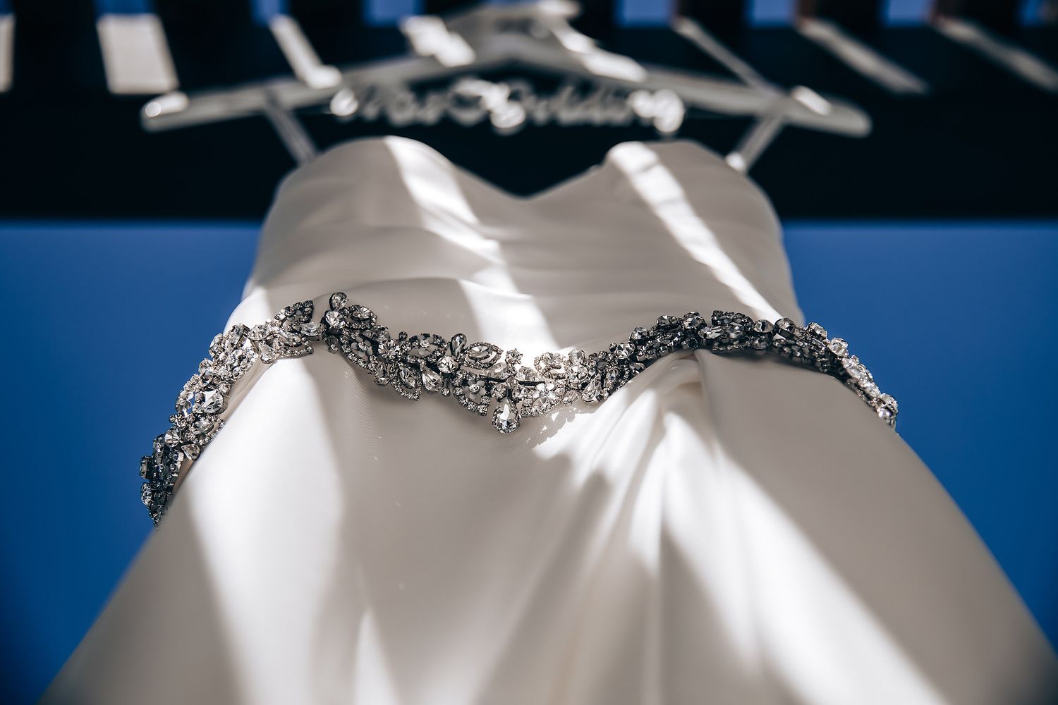 A white satin wedding dress with intricate beaded embellishments hangs elegantly against a blue background.