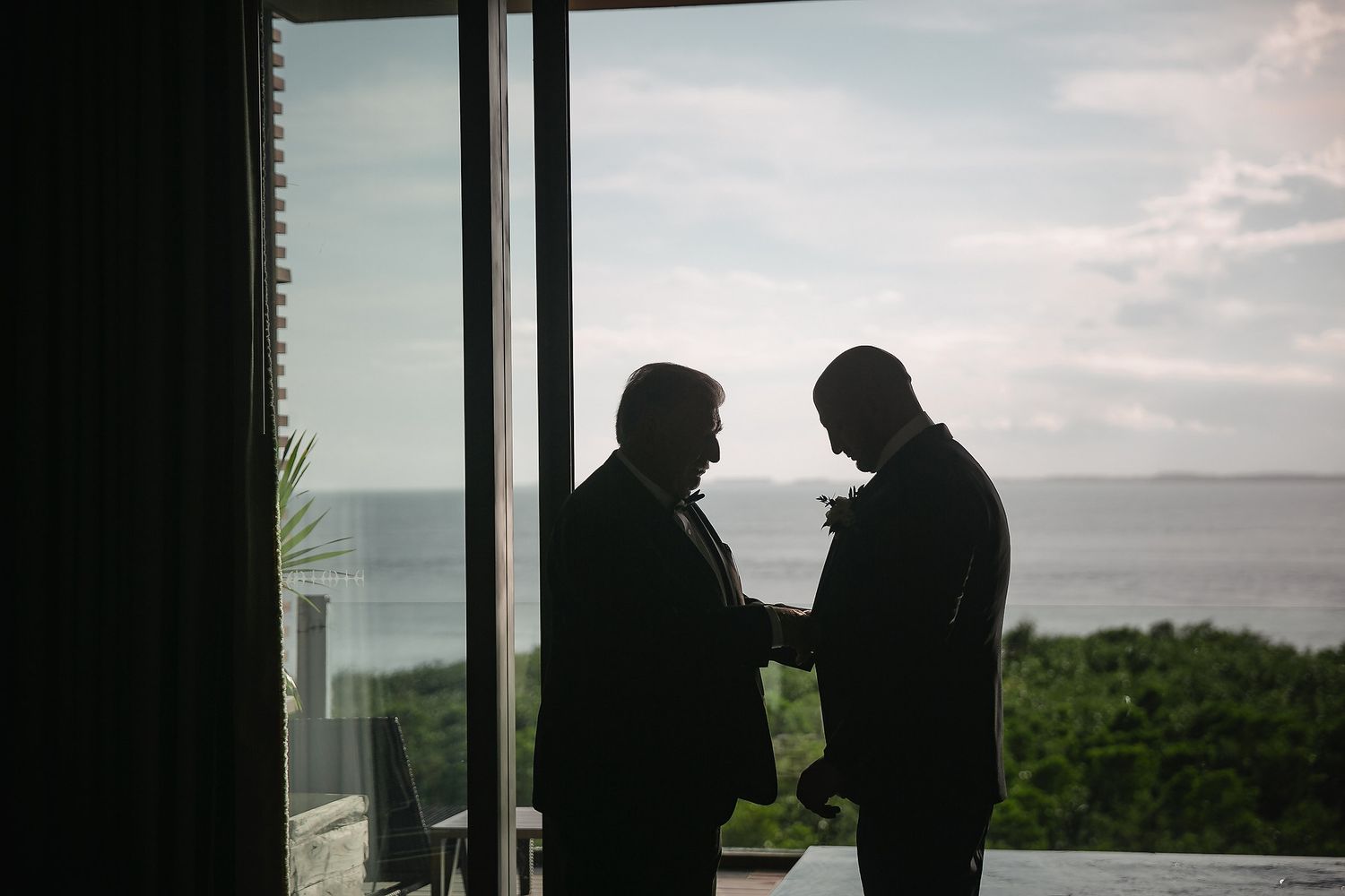 Silhouetted figures stand against large windows overlooking an ocean view at sunset.