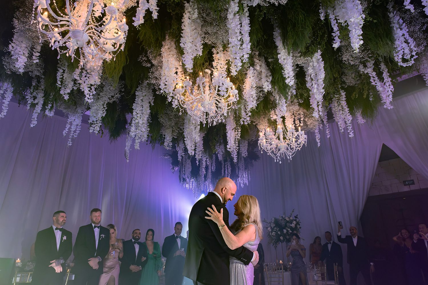 Wedding guests dancing and celebrating on a dance floor under elegant white floral decorations and purple lighting.