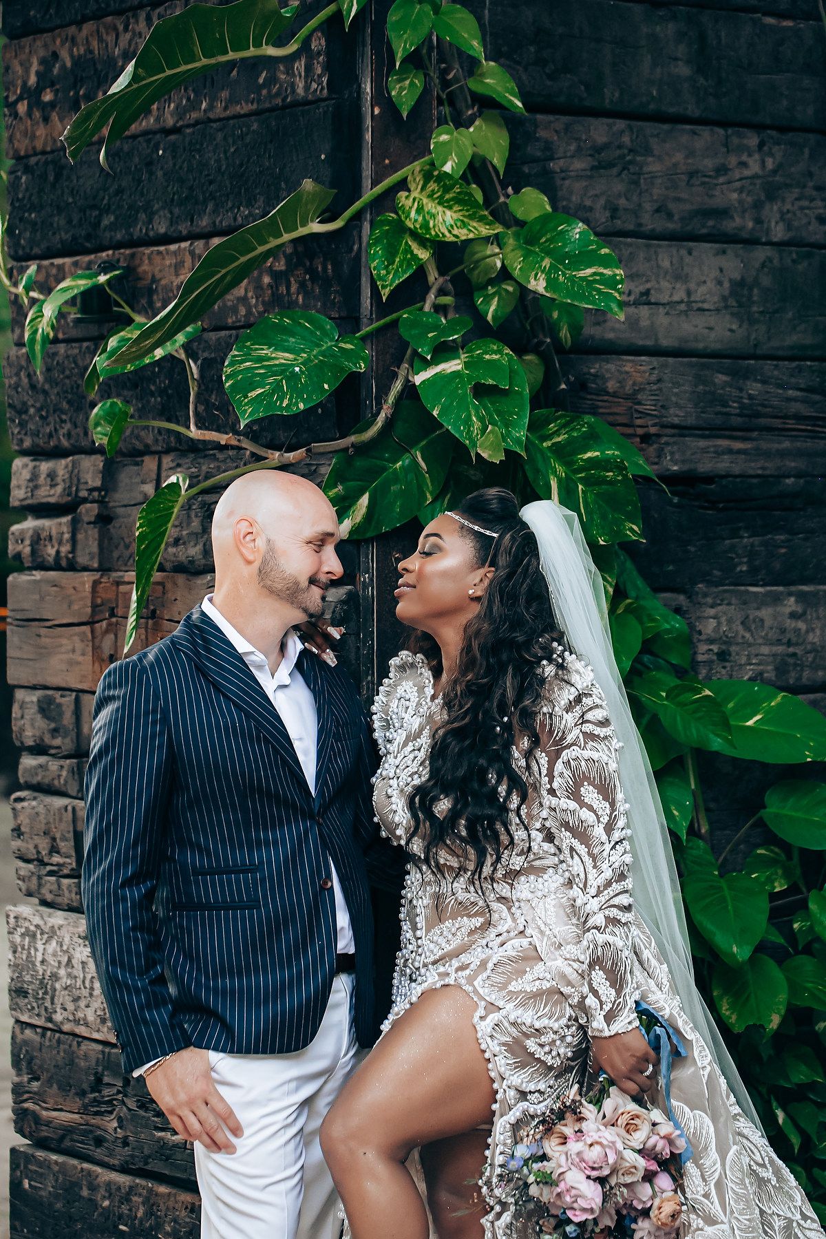 A couple in wedding attire pose against a rustic brick wall with green ivy, sharing an intimate moment together.