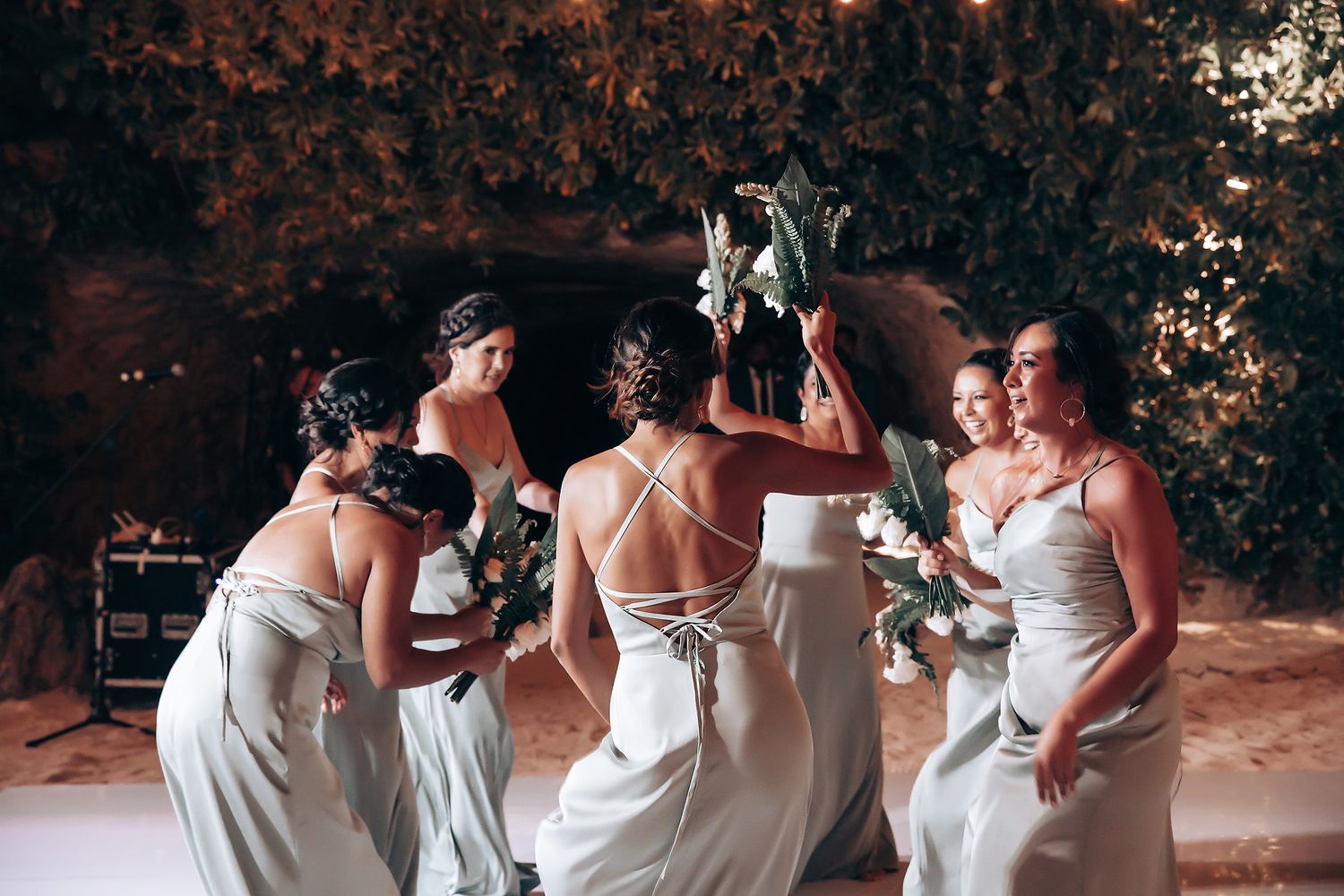Group of bridesmaids in white dresses dancing and celebrating at an outdoor evening reception.