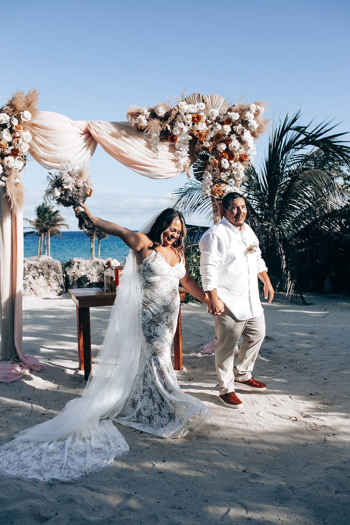 Playful wedding couple dancing and celebrating under a decorated beach arch with ocean views.