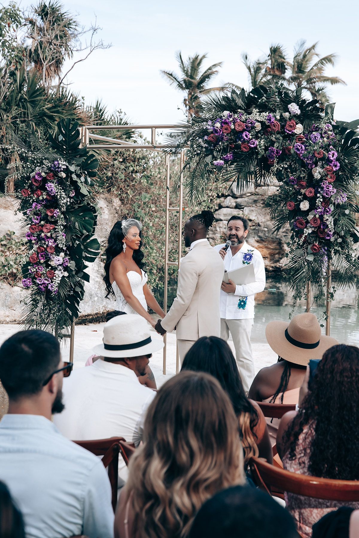 A wedding ceremony takes place outdoors under a floral arch with pink and purple blooms against a tropical backdrop.