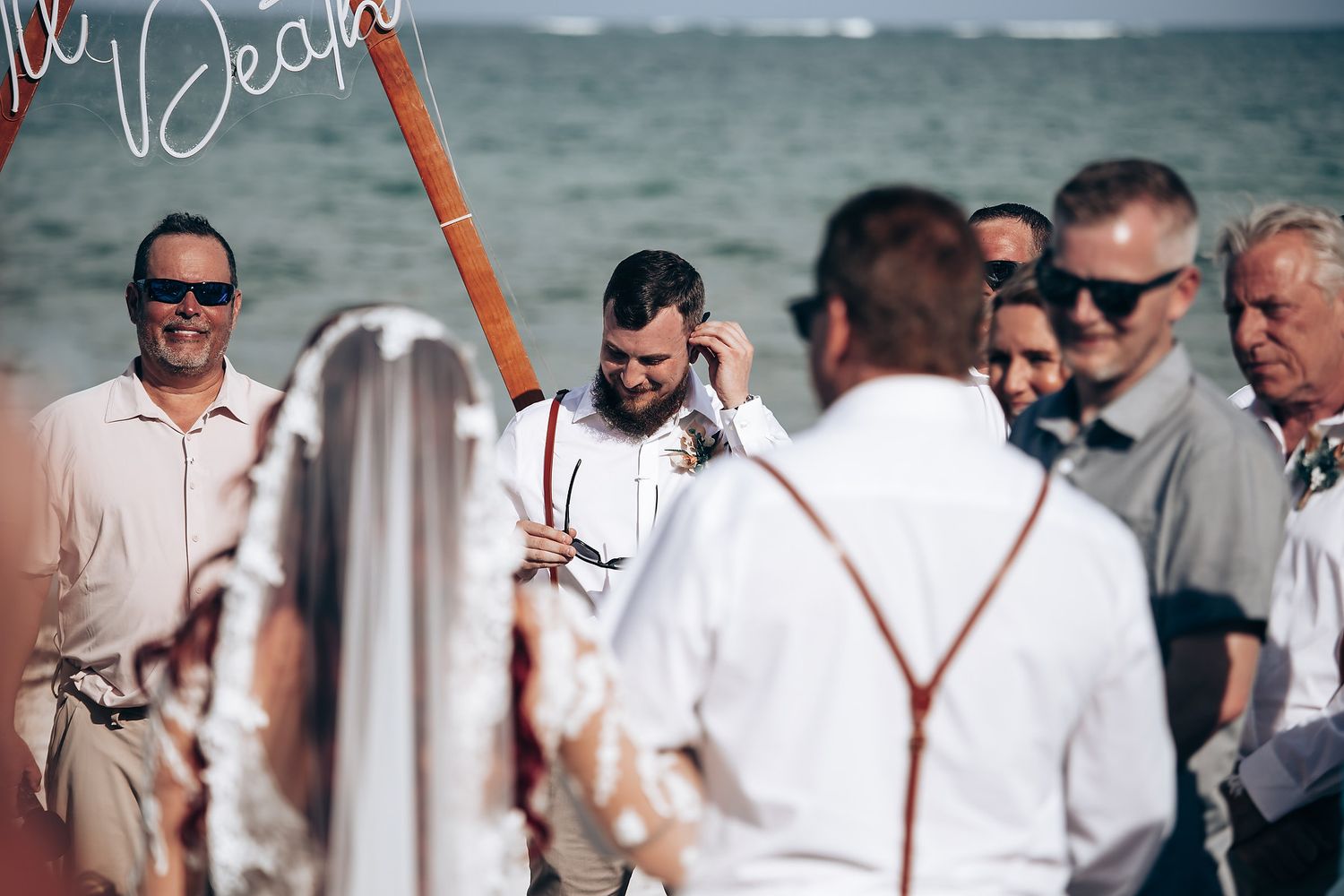 A group of wedding guests in suspenders and formal attire gather on a boat deck with the ocean visible in the background.