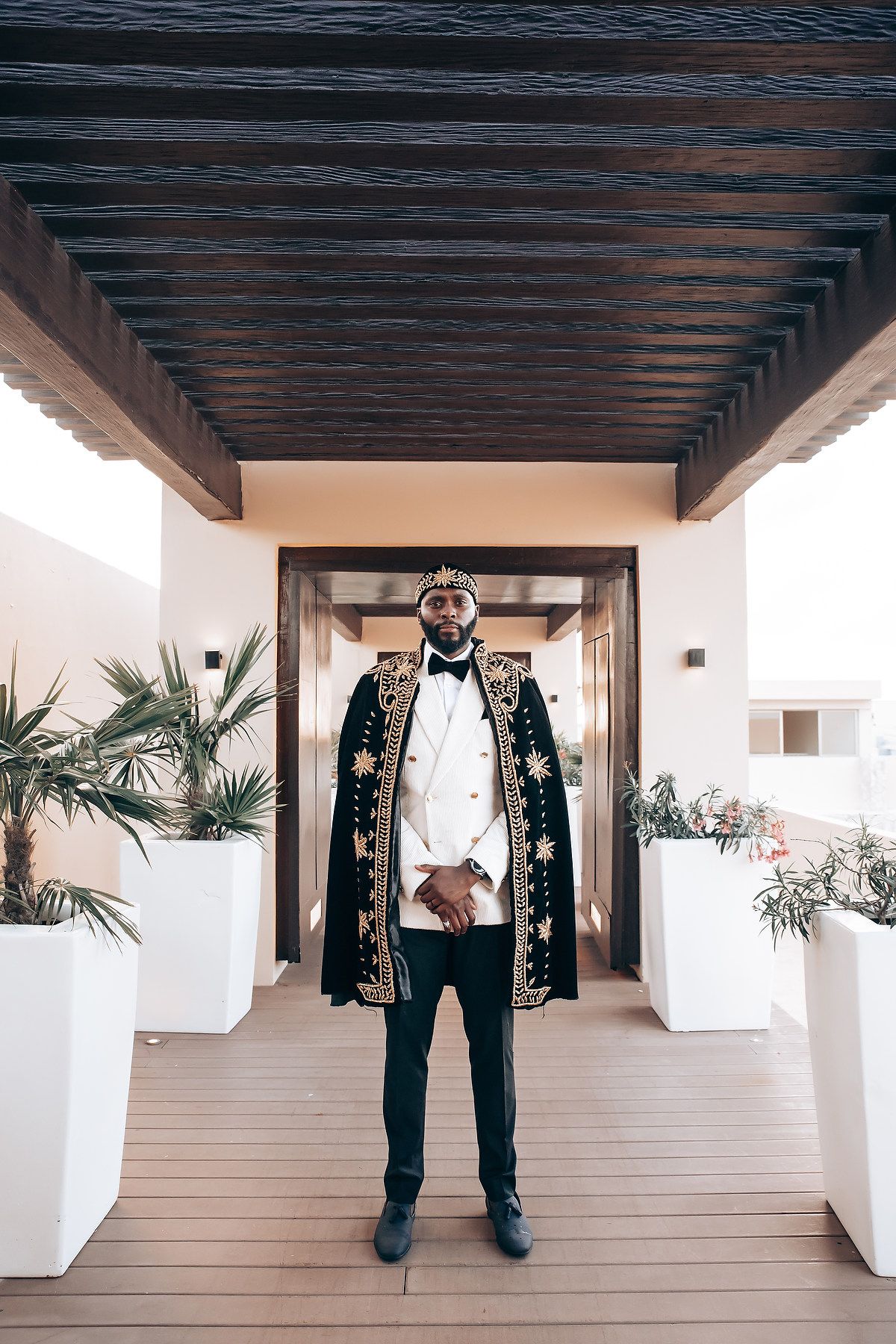 Groom in tuxedo stands in elegant white hallway with wooden beams and tropical plants.
