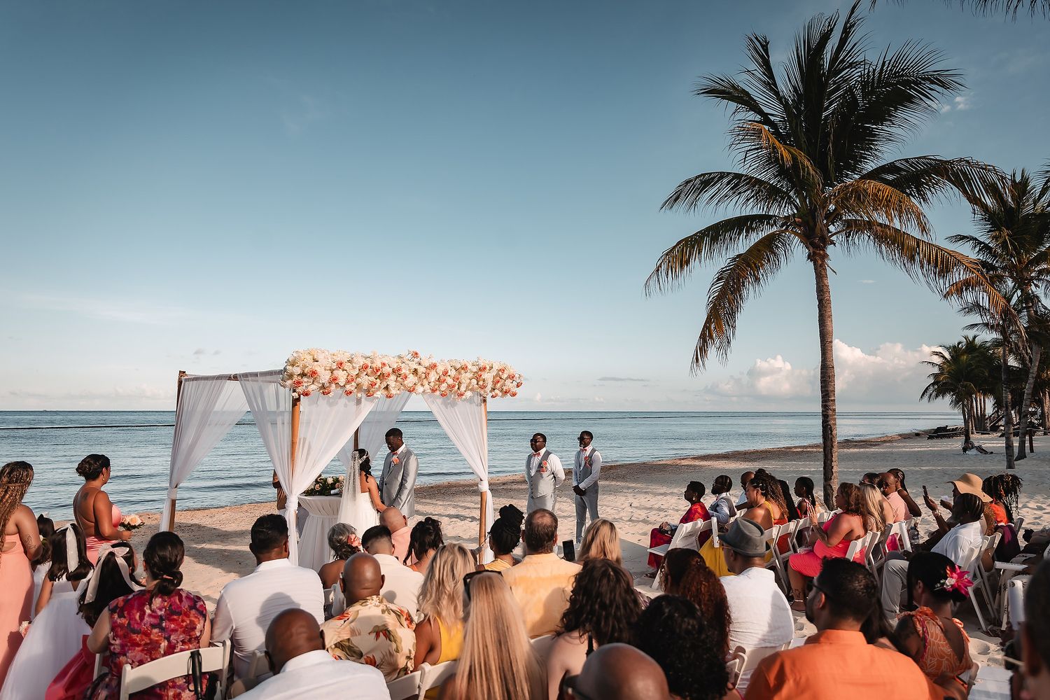 Beachfront wedding ceremony under a floral arch with palm trees and gathered guests.