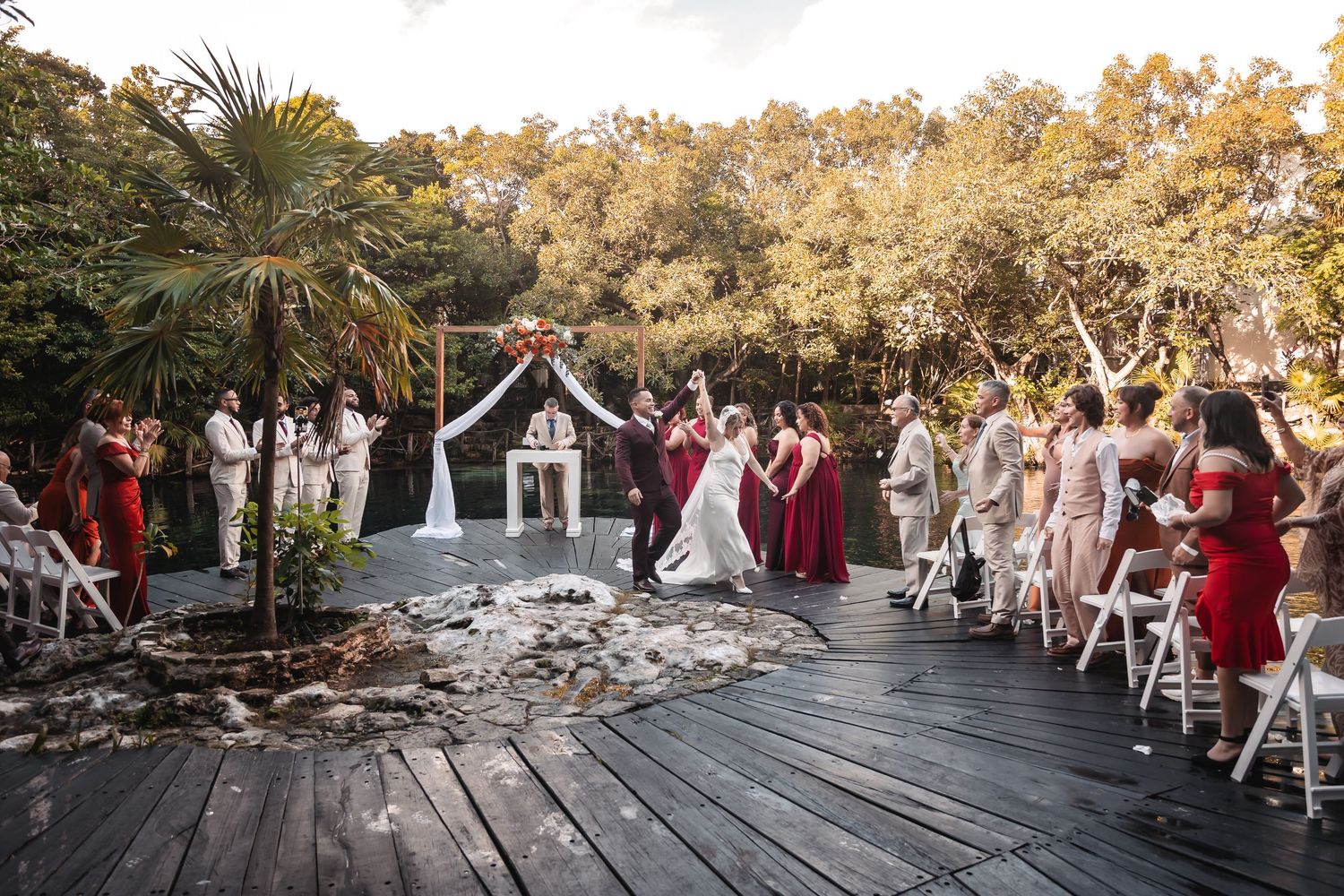 Wedding guests seated in white chairs on wooden deck surrounded by lush greenery.