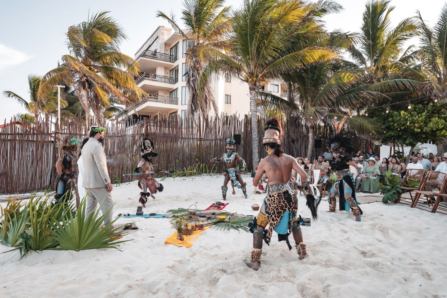 Traditional Mayan dancers in colorful costumes perform on a white sand beach with palm trees and a resort building behind them.