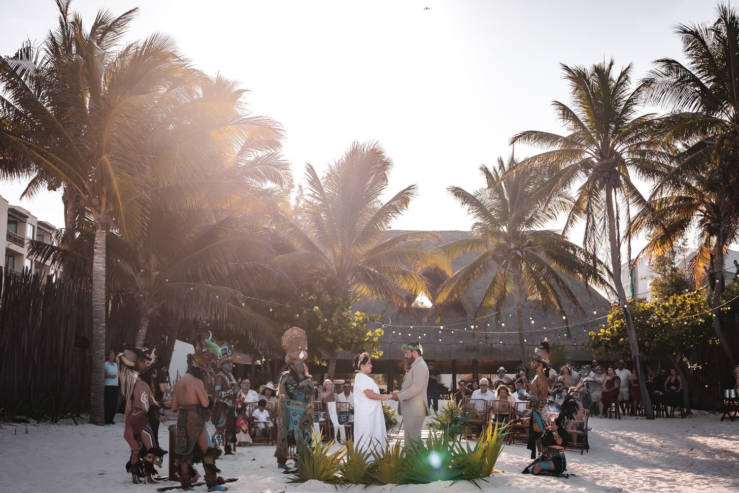 Beach wedding ceremony at sunset with palm trees, guests gathered on sand near a thatched pavilion in a tropical setting.