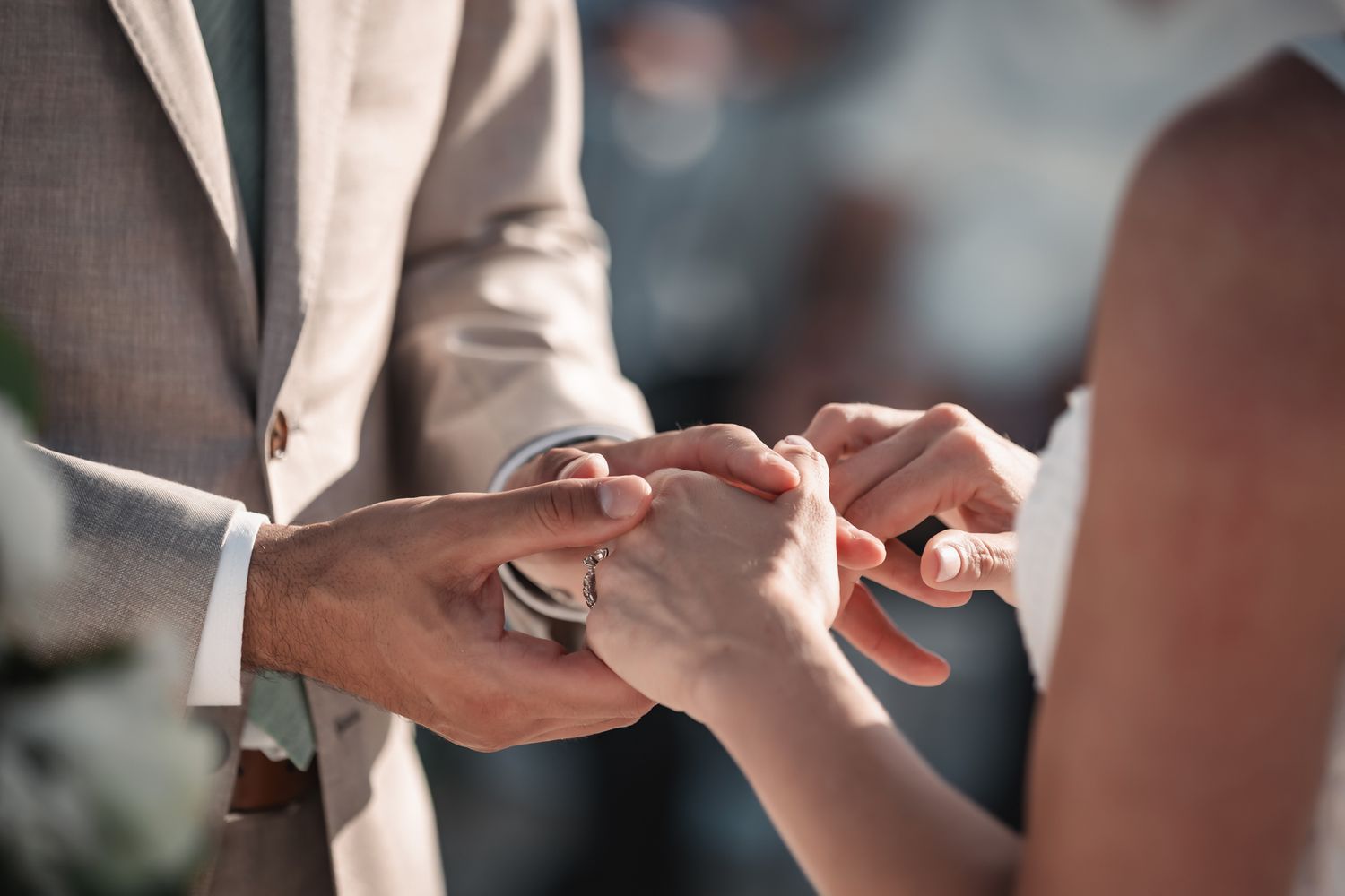 Groom in beige suit exchanging wedding rings with bride during outdoor ceremony.