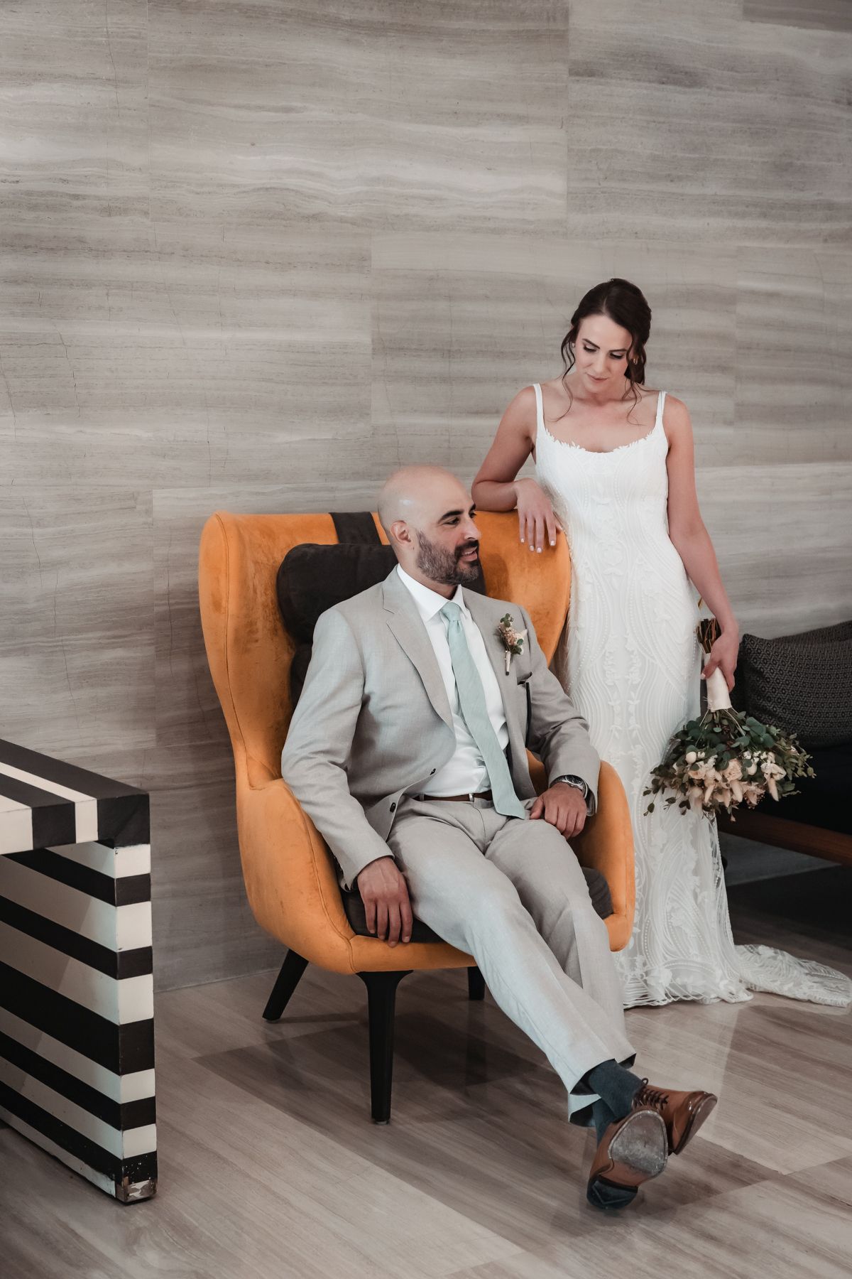 Bride in white gown stands beside groom in tan suit seated in orange chair, holding bouquet in elegant indoor setting.