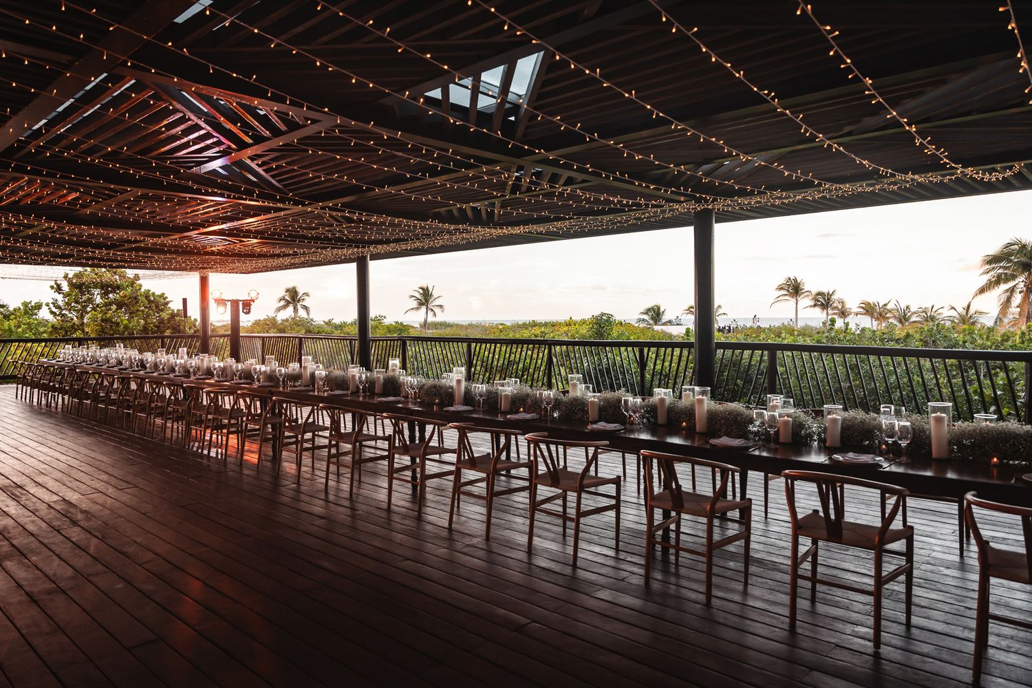Elegant outdoor wedding reception with long dining tables, string lights, and tropical views at golden hour.