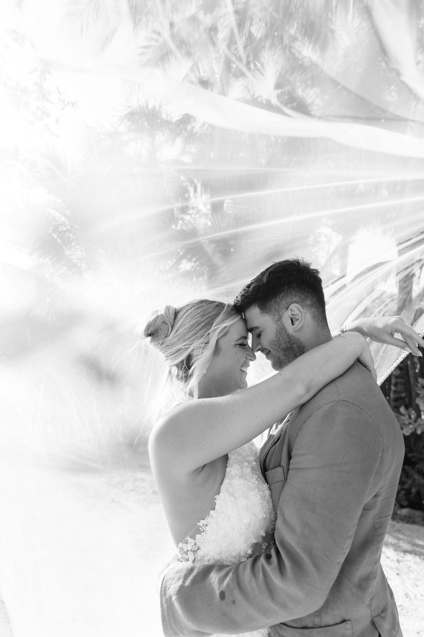 Black and white photo of bride and groom embracing under flowing veil among palm trees.