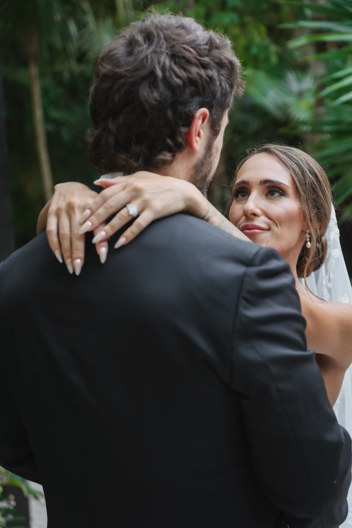 Bride gazes lovingly at groom during outdoor wedding dance, wearing veil and diamond earrings amid lush greenery.
