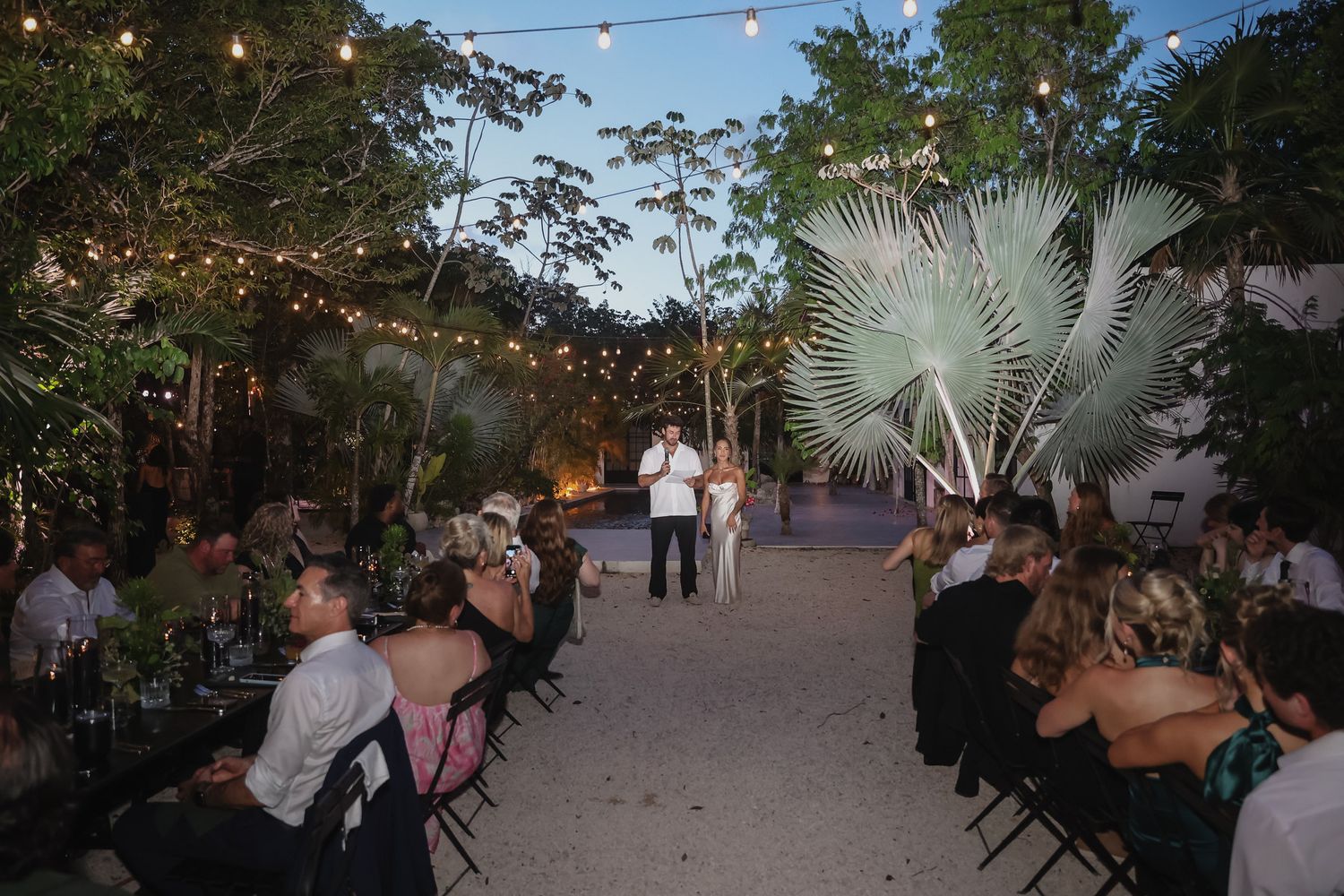 Outdoor wedding reception at night with string lights, tropical palms, and guests seated on both sides of a sandy aisle.
