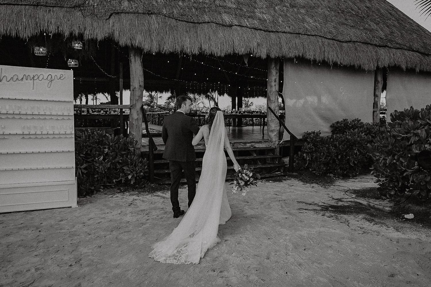 Black and white photo of bride and groom walking toward a thatched beach bar on their wedding day.