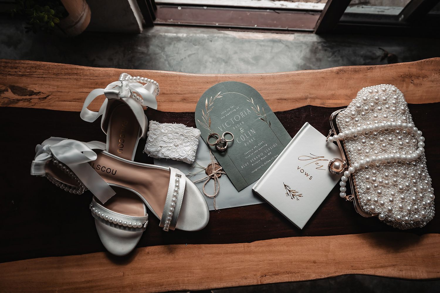 Elegant wedding flat lay featuring white pearl heels, pearl clutch, sage green invitation suite, rings, and delicate accessories on wood.