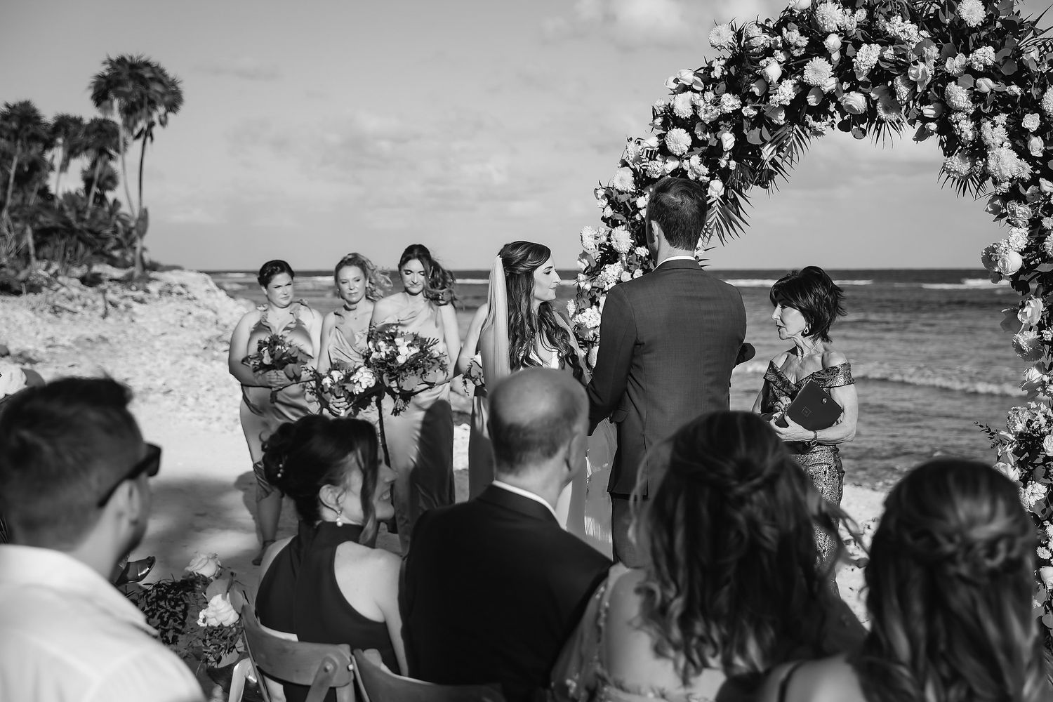 Black and white photo of bridesmaids and groom at a beach wedding ceremony with a lush floral arch backdrop.