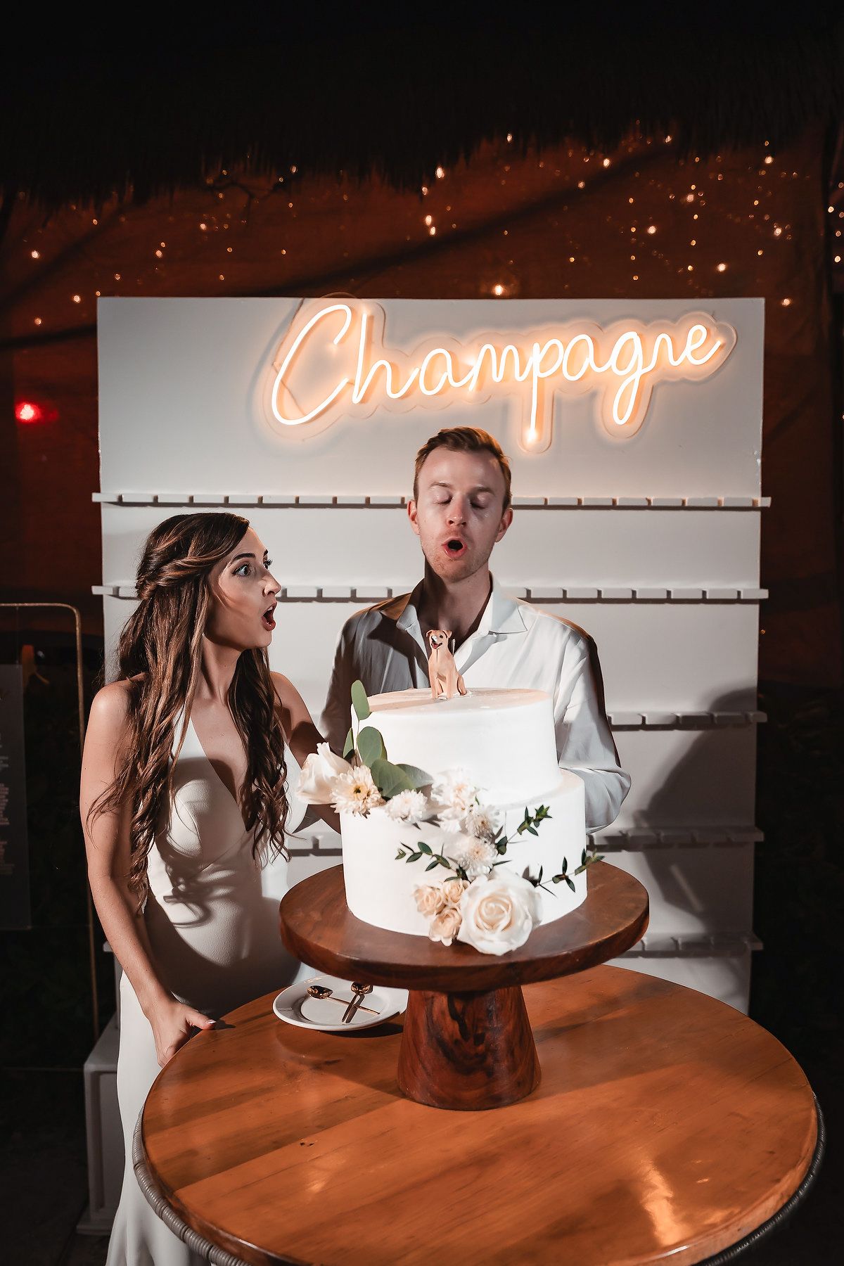 Bride and groom blowing out candle on white floral wedding cake under a neon Champagne sign.