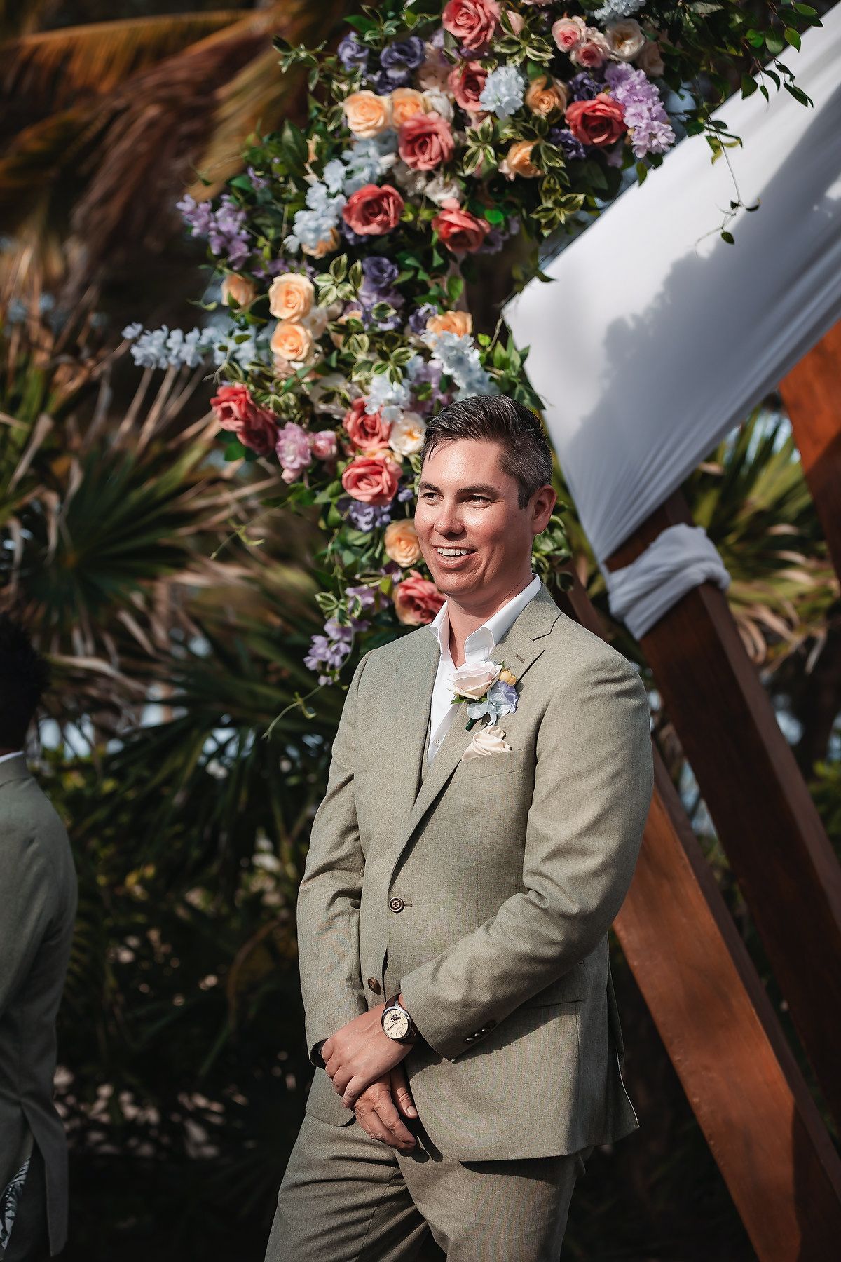 Smiling groom in sage green suit with boutonniere stands before colorful floral arch at outdoor wedding ceremony.