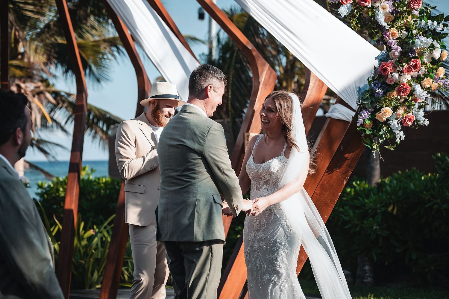 Couple exchanging vows at outdoor beach wedding ceremony with floral arch and officiant in tropical setting.