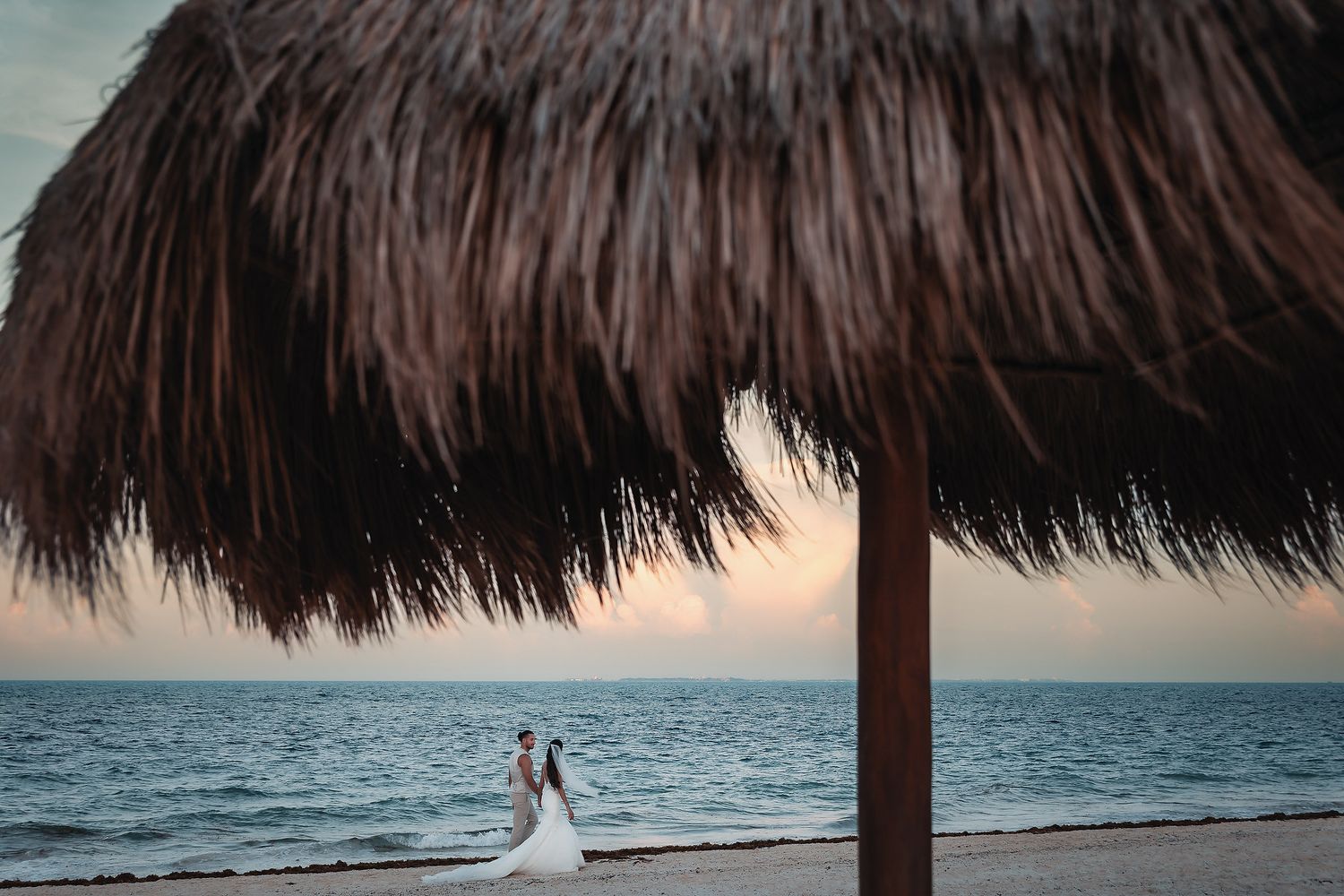 Bride and groom embrace on a tropical beach at sunset beneath a thatched palapa umbrella.