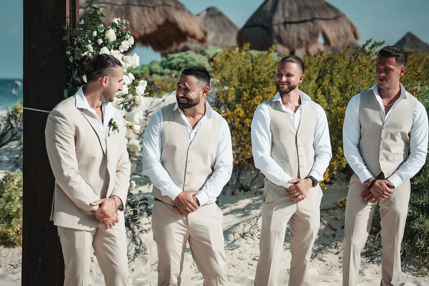 Four groomsmen in matching beige linen suits stand together on a beach wedding ceremony with tropical decor.