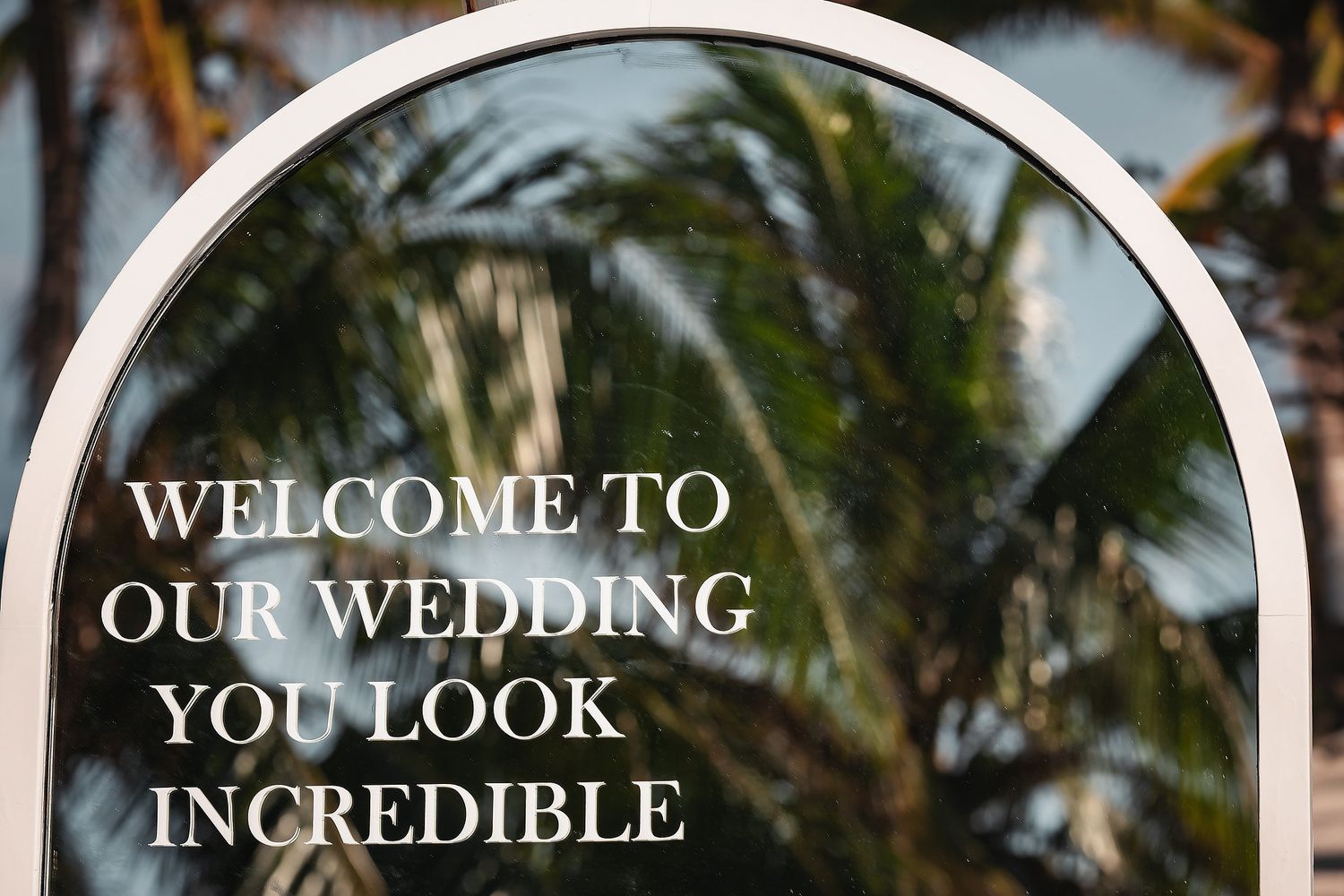 Elegant arched mirror wedding welcome sign reading You Look Incredible, framed by lush tropical palm trees.