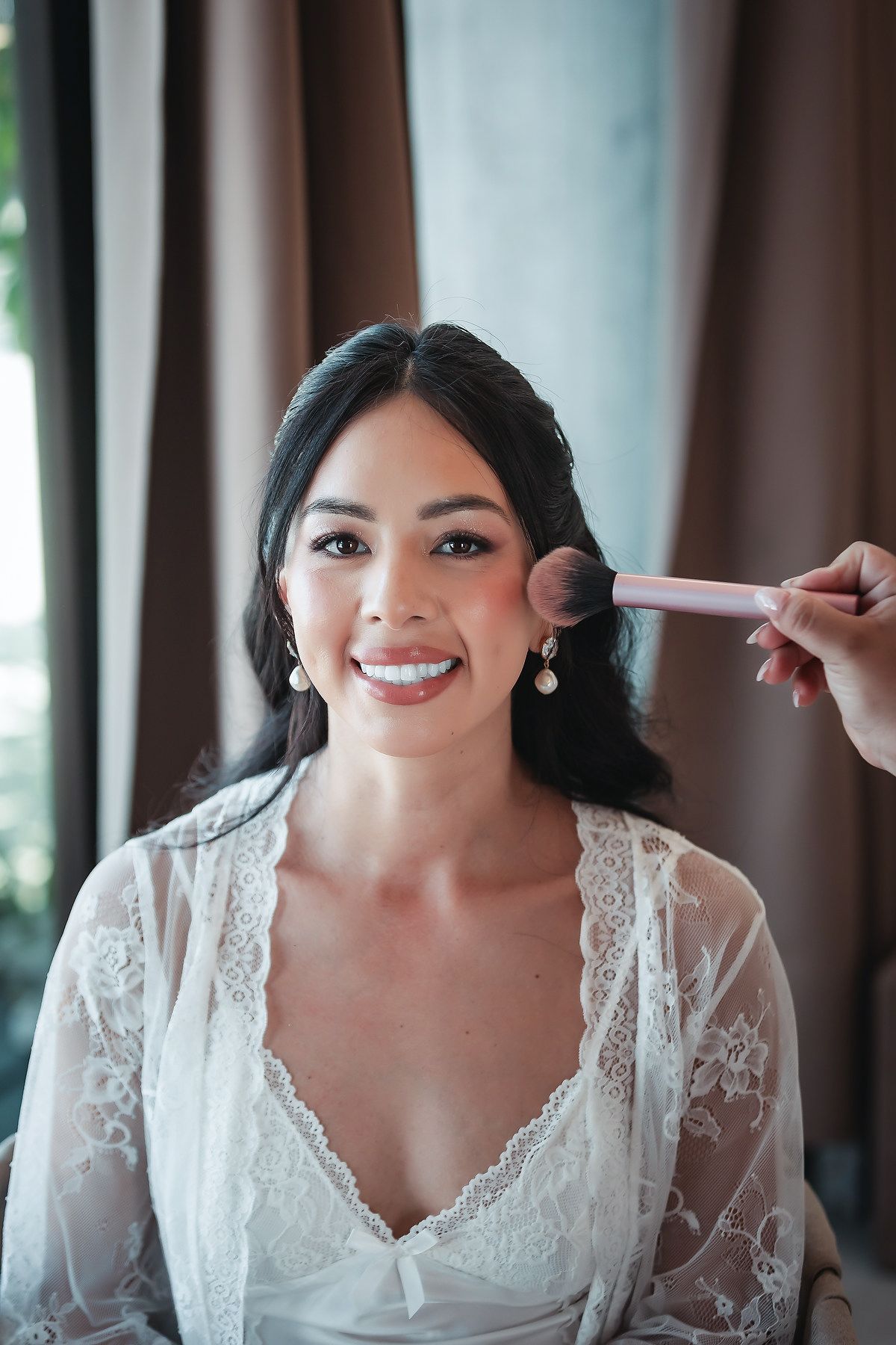 A smiling bride in a white lace robe gets blush applied with a pink makeup brush during her wedding day preparations.
