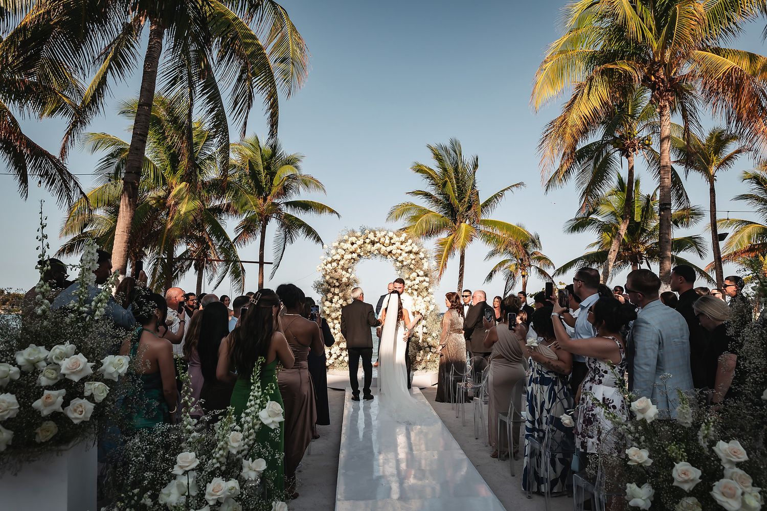 Guests stand for couple at tropical beach wedding with circular floral arch and palm trees.