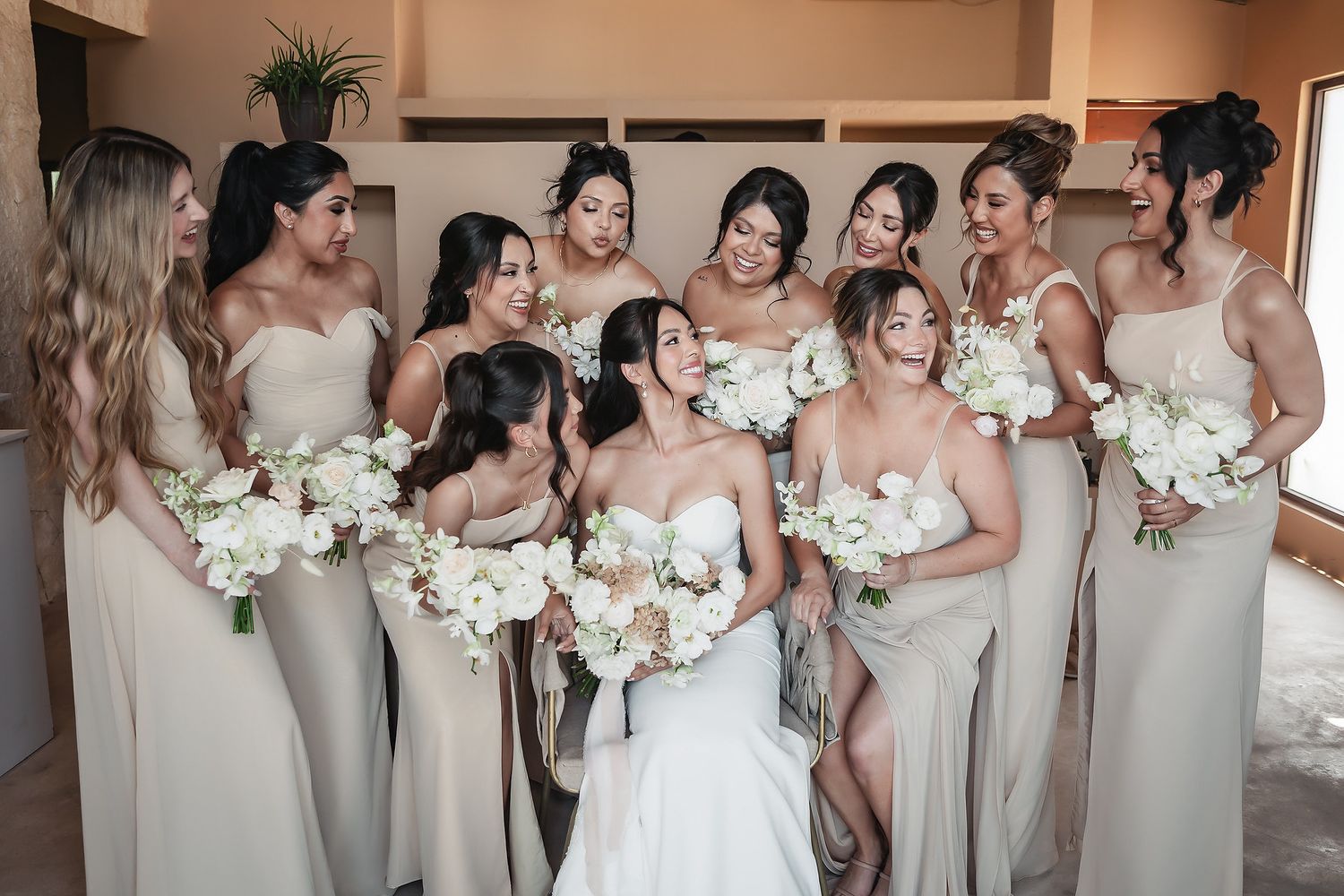 Bride surrounded by bridesmaids in champagne gowns holding white floral bouquets indoors.