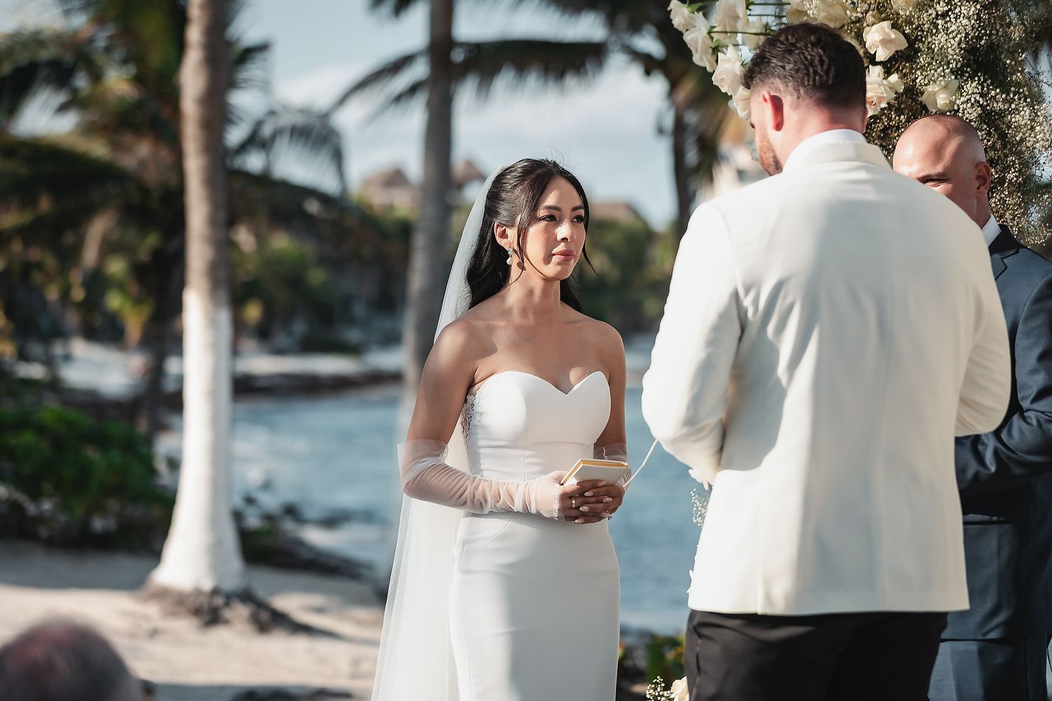 Bride smiling at groom during outdoor beach wedding ceremony with palm trees and ocean in background.