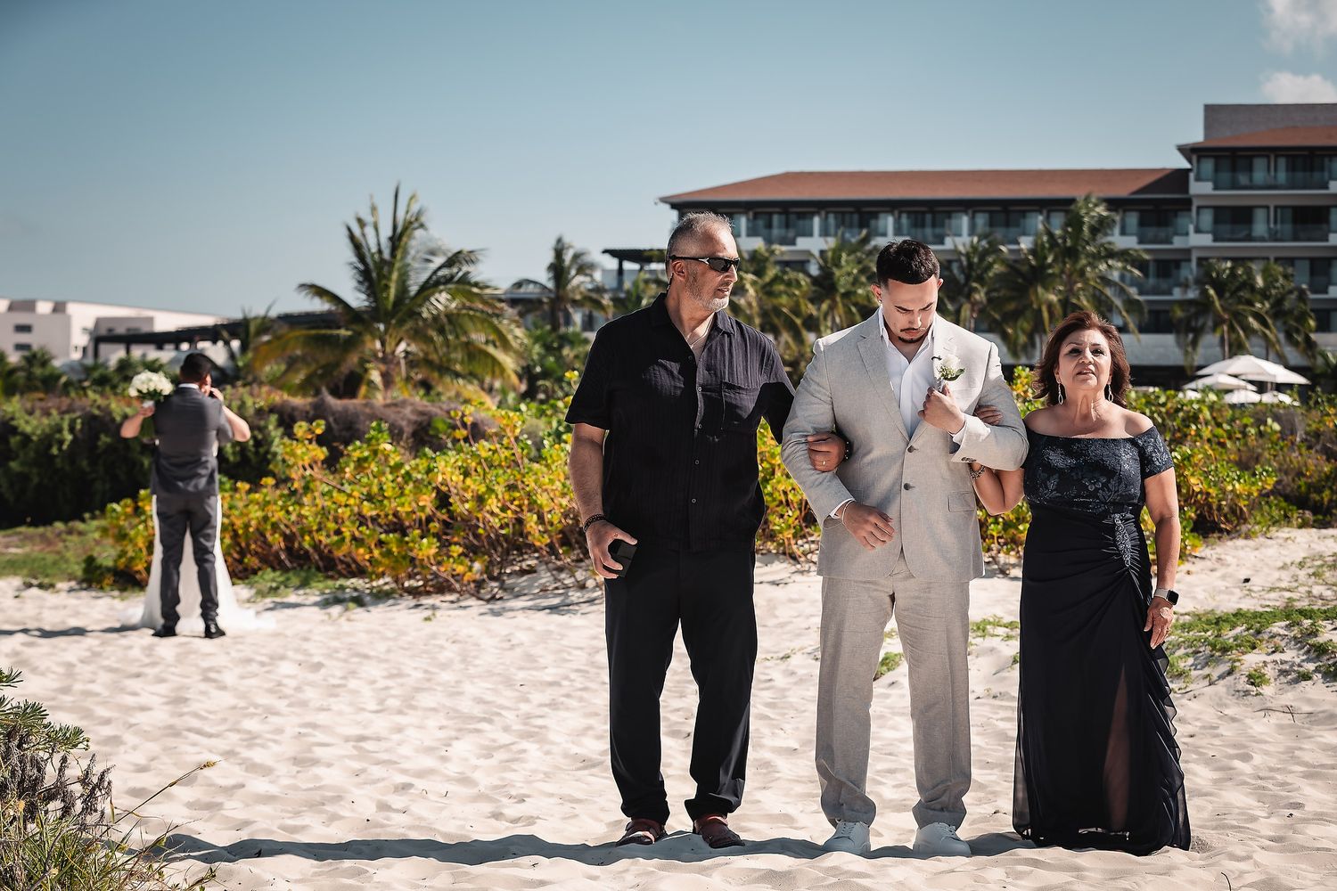Groom in gray suit walks with parents on sandy beach with resort in background on wedding day.