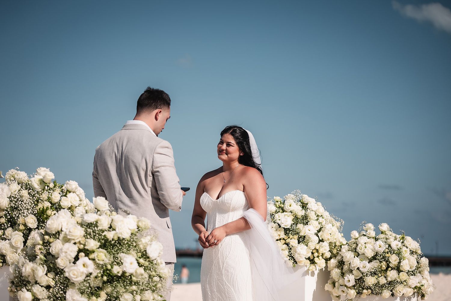 Couple exchanging vows at a beachfront wedding ceremony surrounded by lush white floral arrangements under a clear blue sky.
