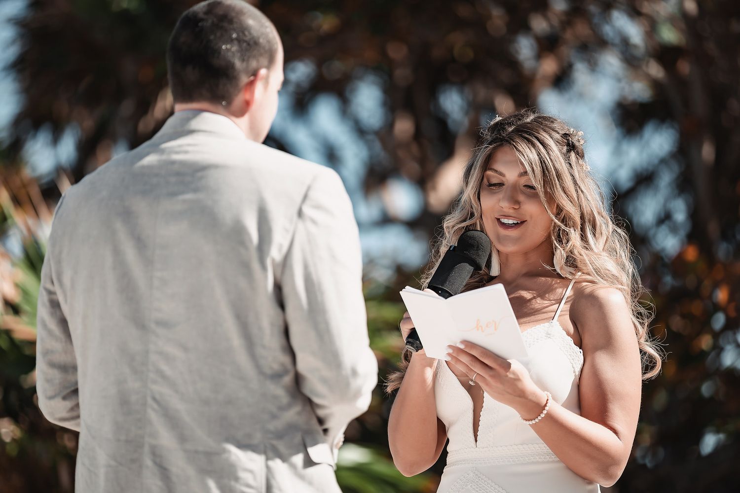 Bride smiling while reading wedding vows from a card during an outdoor ceremony, wearing a white dress.