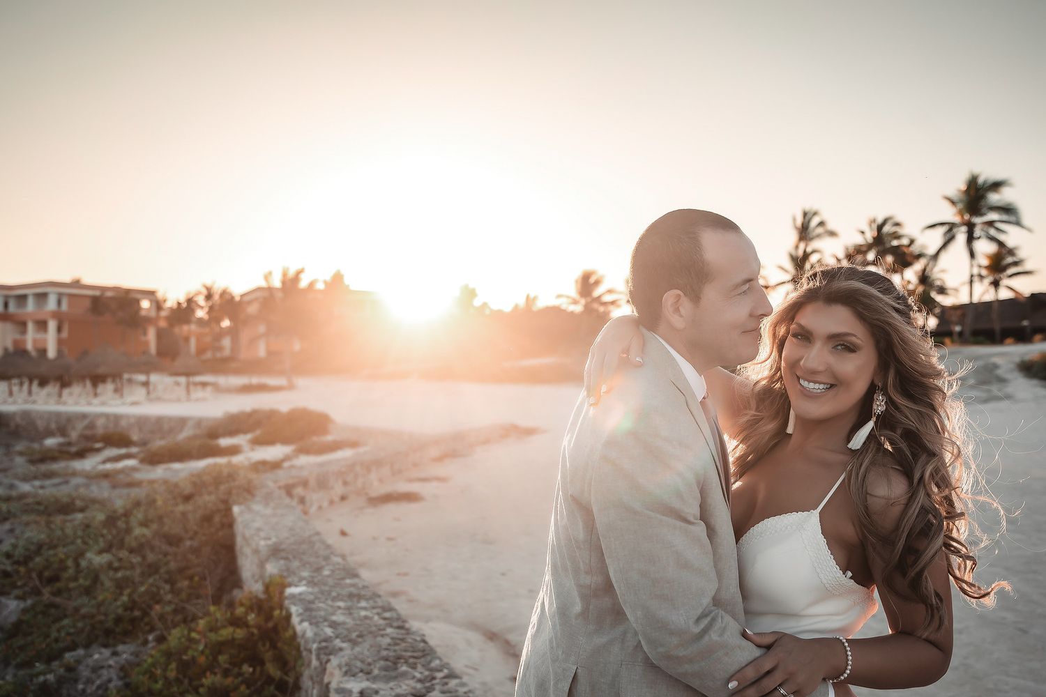 Bride and groom embrace on a sandy beach at golden sunset with palm trees and resort in the background.