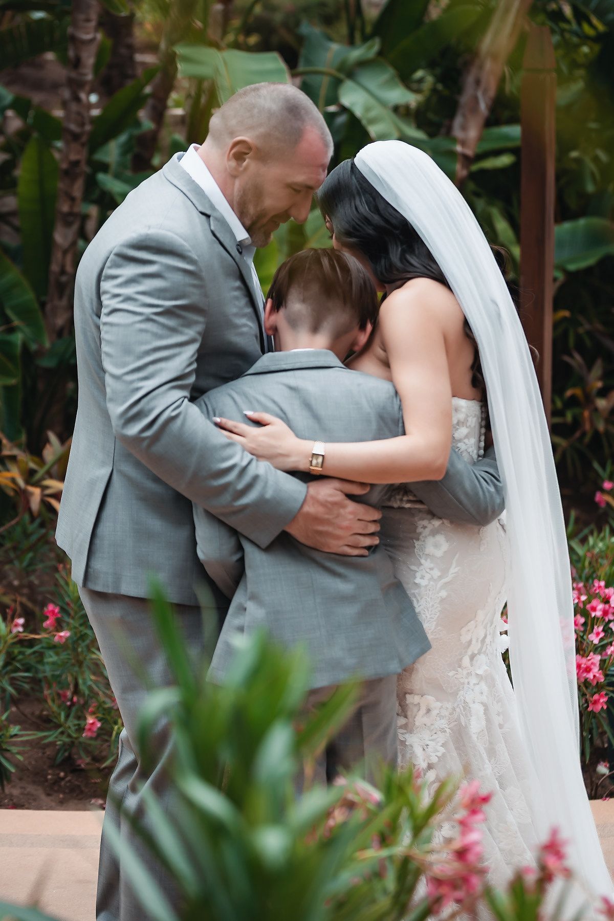 Bride and groom in gray suits share emotional family hug with child during outdoor wedding ceremony surrounded by tropical greenery.