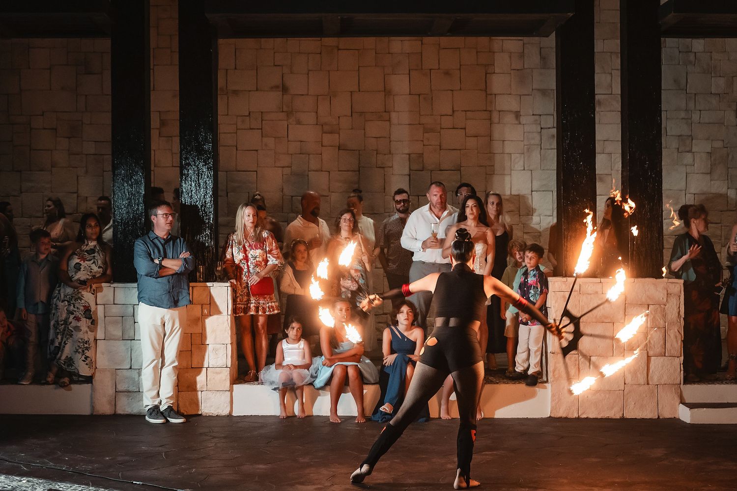 Fire dancer performing with spinning flames for a crowd of spectators at an outdoor stone venue at night.